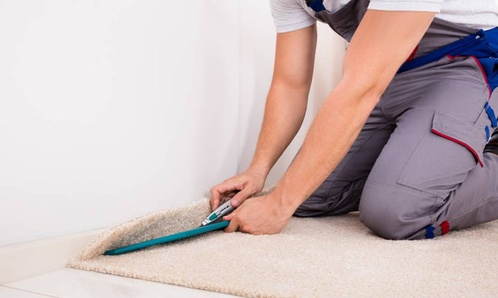 A professional carpet installer kneels near a baseboard to trim the edge of new tan carpeting using a utility knife.