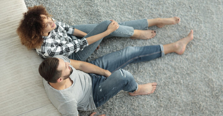 A smiling young couple lean against their sofa and sit on the gray carpet in the living room of their home.