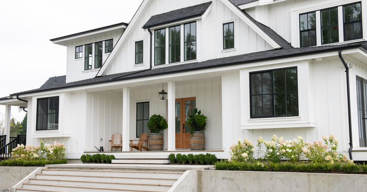 A modern farmhouse with white siding and black window trim features concrete steps and landscaping near the front porch.