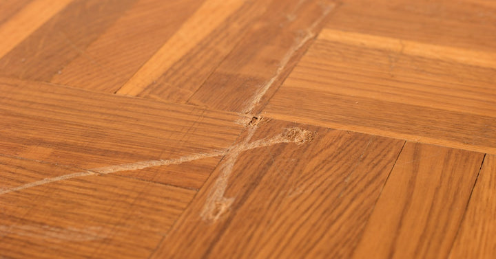 A deep x-shaped scratch with visible indentations cuts across a hardwood floor, with one panel corner missing a chip.
