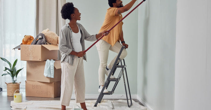 A man and woman paint a room in their home. The man stands on a ladder to paint and the woman stands on the floor.