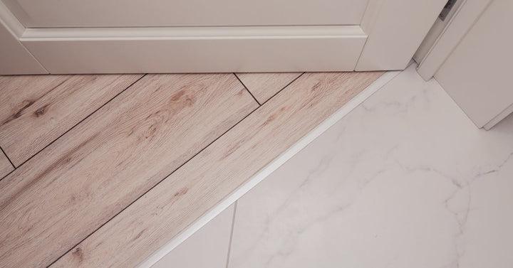 A close-up of a floor transition shows light wood planks meeting white marble tiles, divided by a silver strip.